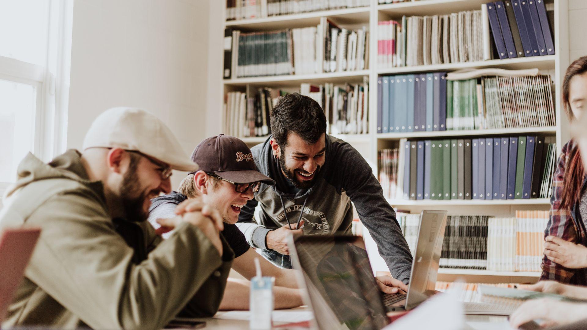 Four individuals in front of a white bookshelf filled with books and binders. Three men are in the foreground, two of whom are laughing and leaning towards a laptop on a table. The third man in a cap is looking down. To the right, a part of another individual is visible, focused on a separate laptop