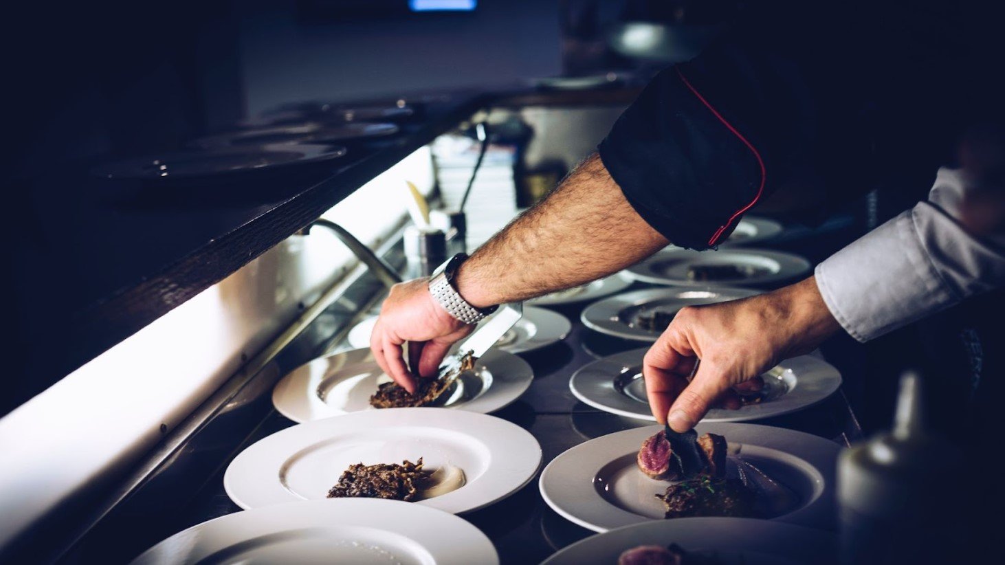 Close-up of a chef's hands meticulously placing food on white plates in a dimly lit kitchen